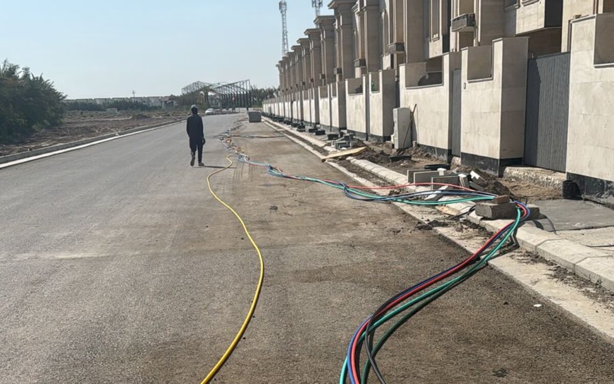 Bundle of coloured conduits routed along the kerb beside a row of newly built units at Al-Khaleej Complex, with a worker on the access road